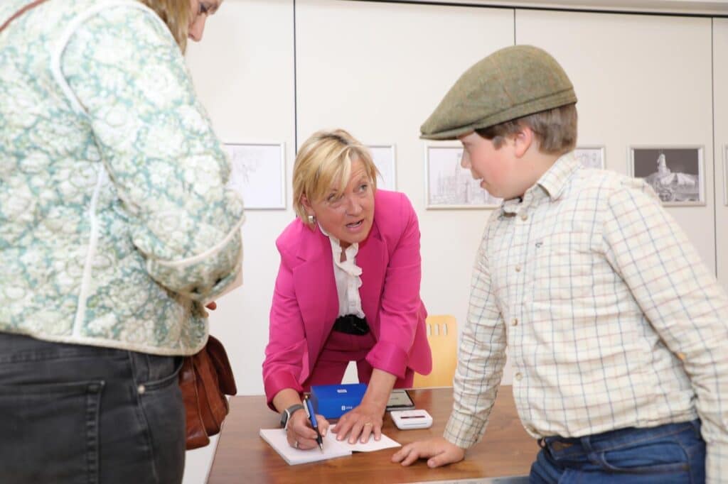 A blonde lady in a pink jacket at a book signing talking to a young boy