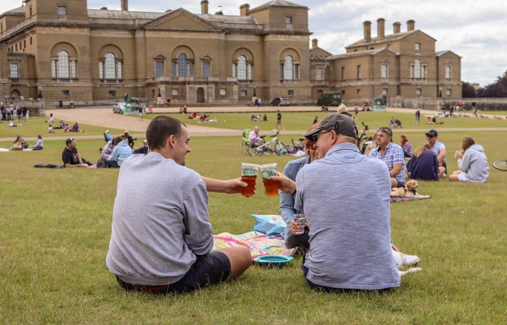 Two men in blue tops cheers the beers as they sit on the grass in front of the North side of Holkham Hall