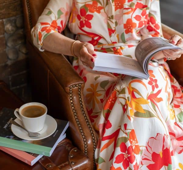 A lady in a colourful floral dress reads a book, sitting in a leather armchair. A cup of coffee stands on a stack of books beside her.