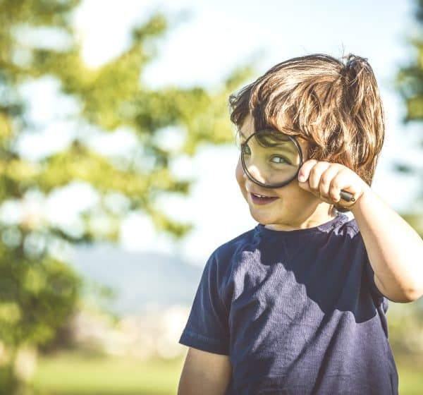 A small boy stands outside looking through a magnifying glass