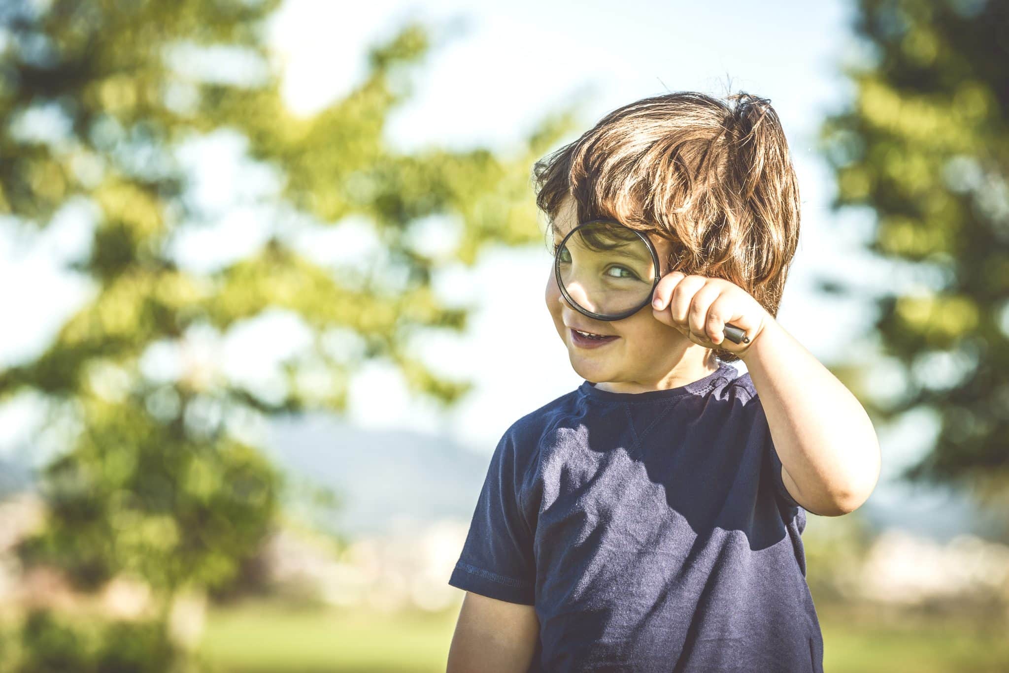 A small boy stands outside looking through a magnifying glass