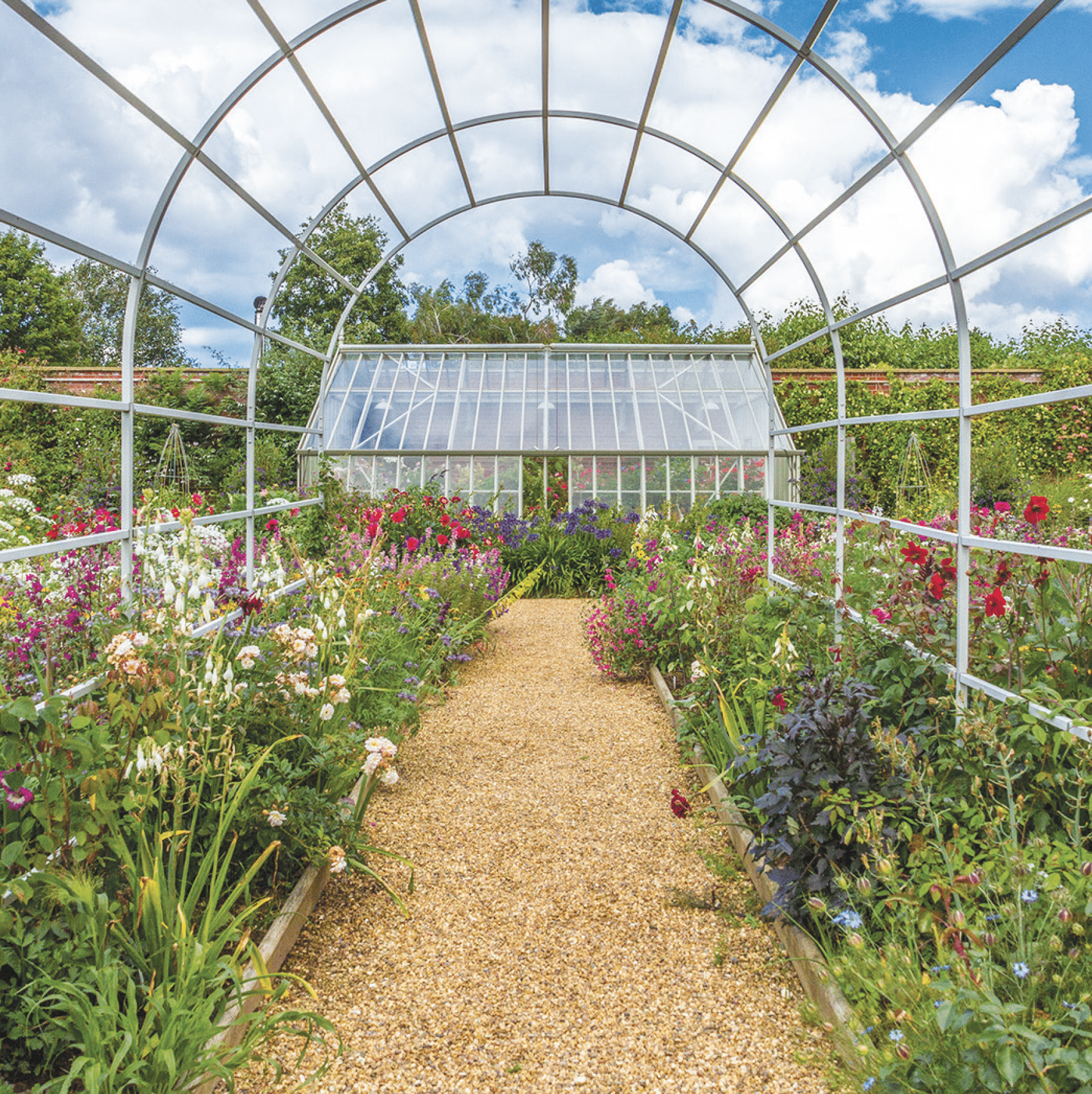 East Ruston Old Vicarage Diamond Jubilee garden, looking towards a glasshouse, with various colourful flowers growing to the sides of a gravel path.