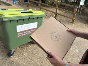 A white person holding a brown compostable pizza box in front of a custom green compost bin. 