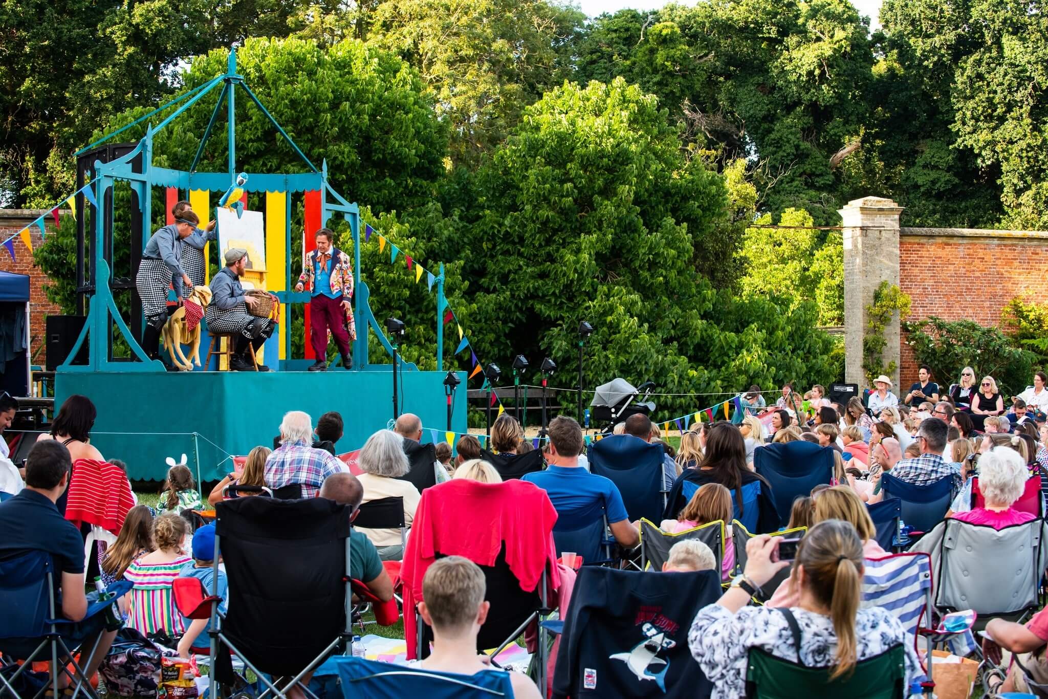 An audience watch a show on a small wooden stage in the open air