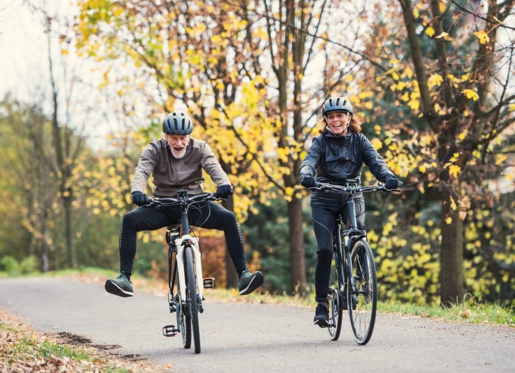 Two adults cycle on an autumn day. The man has his legs out and a playful expression while the woman laughs