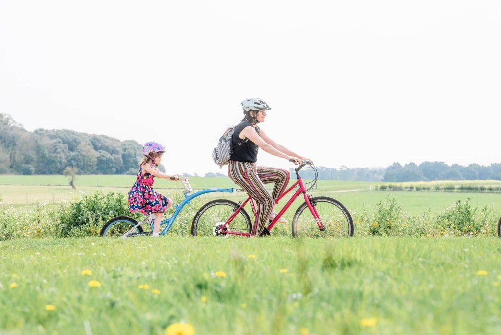 A mum cycles through a Holkham field, pulling her daughter on a tag bike