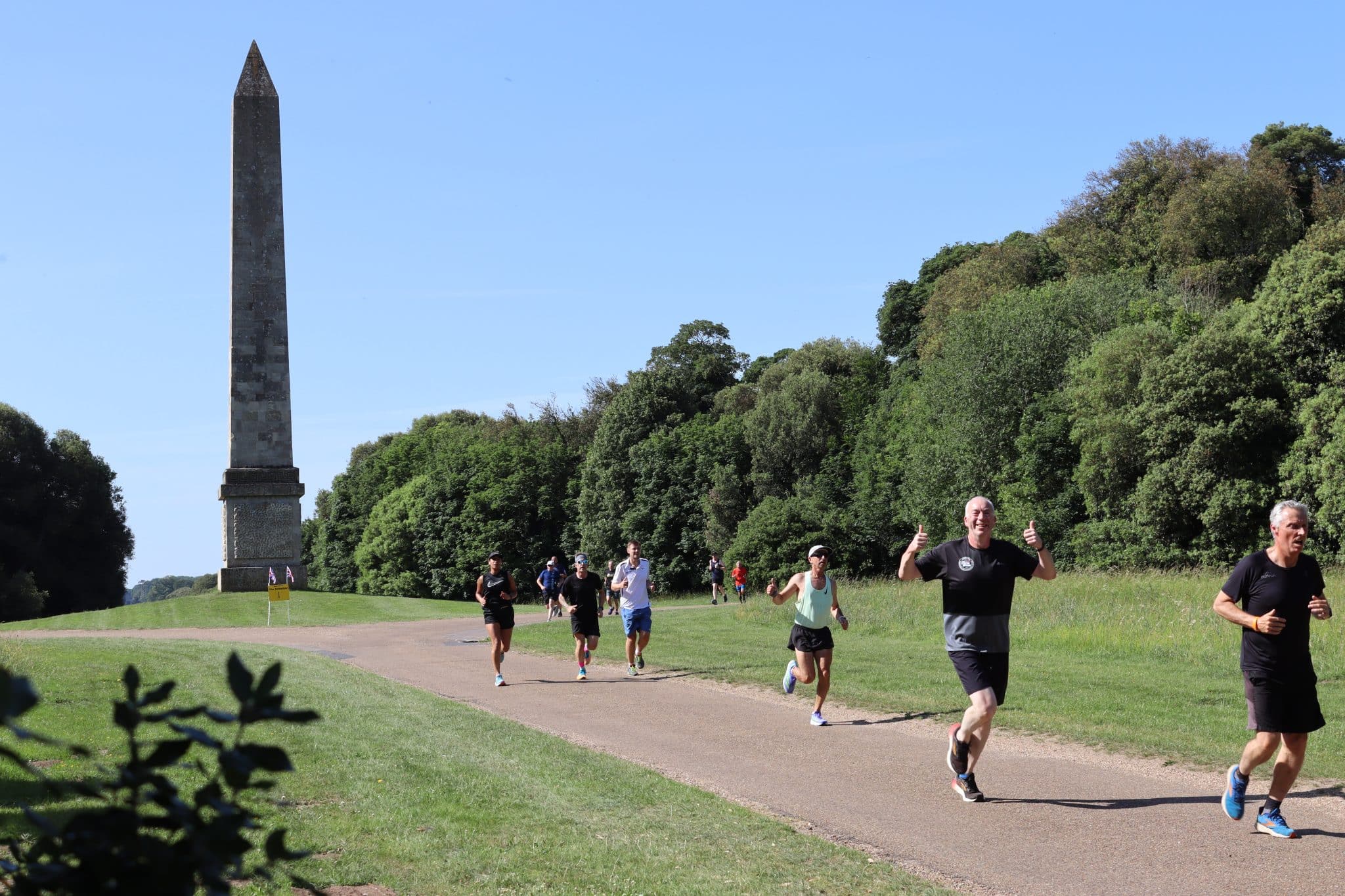 Runners run along a concrete path in the parkland of Holkham Park, with the Obelisk in the background