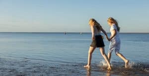 Two girls in summer clothing paddle in the sea, holding hands.