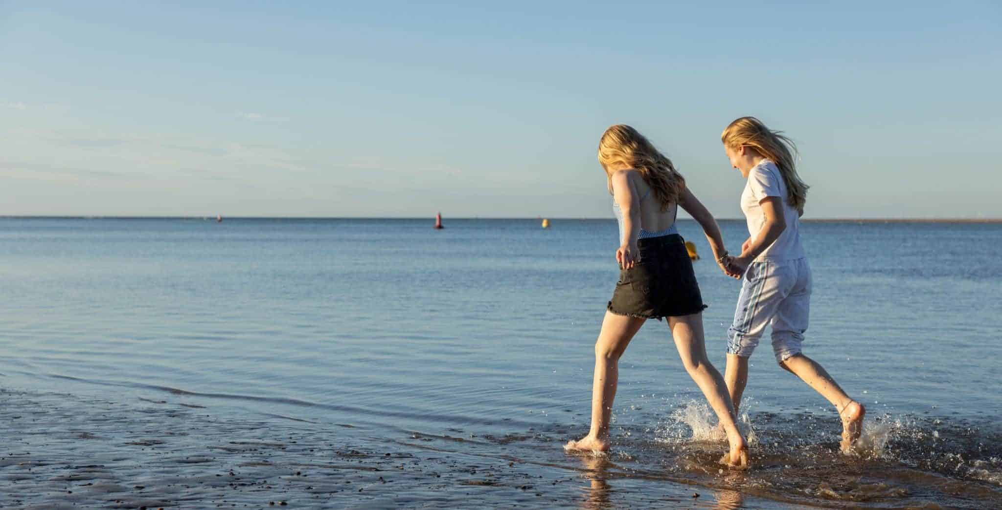 Two girls in summer clothing paddle in the sea, holding hands.