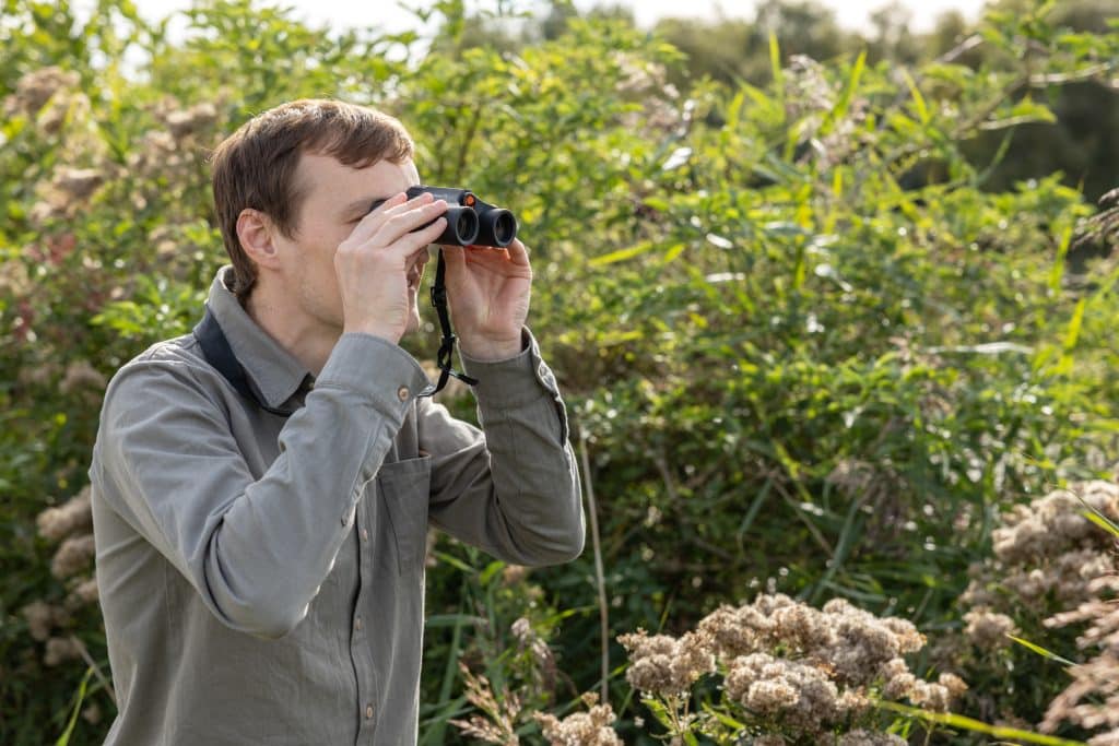 A man looks through binoculars surrounded by trees in North Norfolk