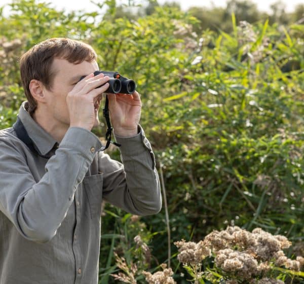 A man looks through binoculars surrounded by trees in North Norfolk
