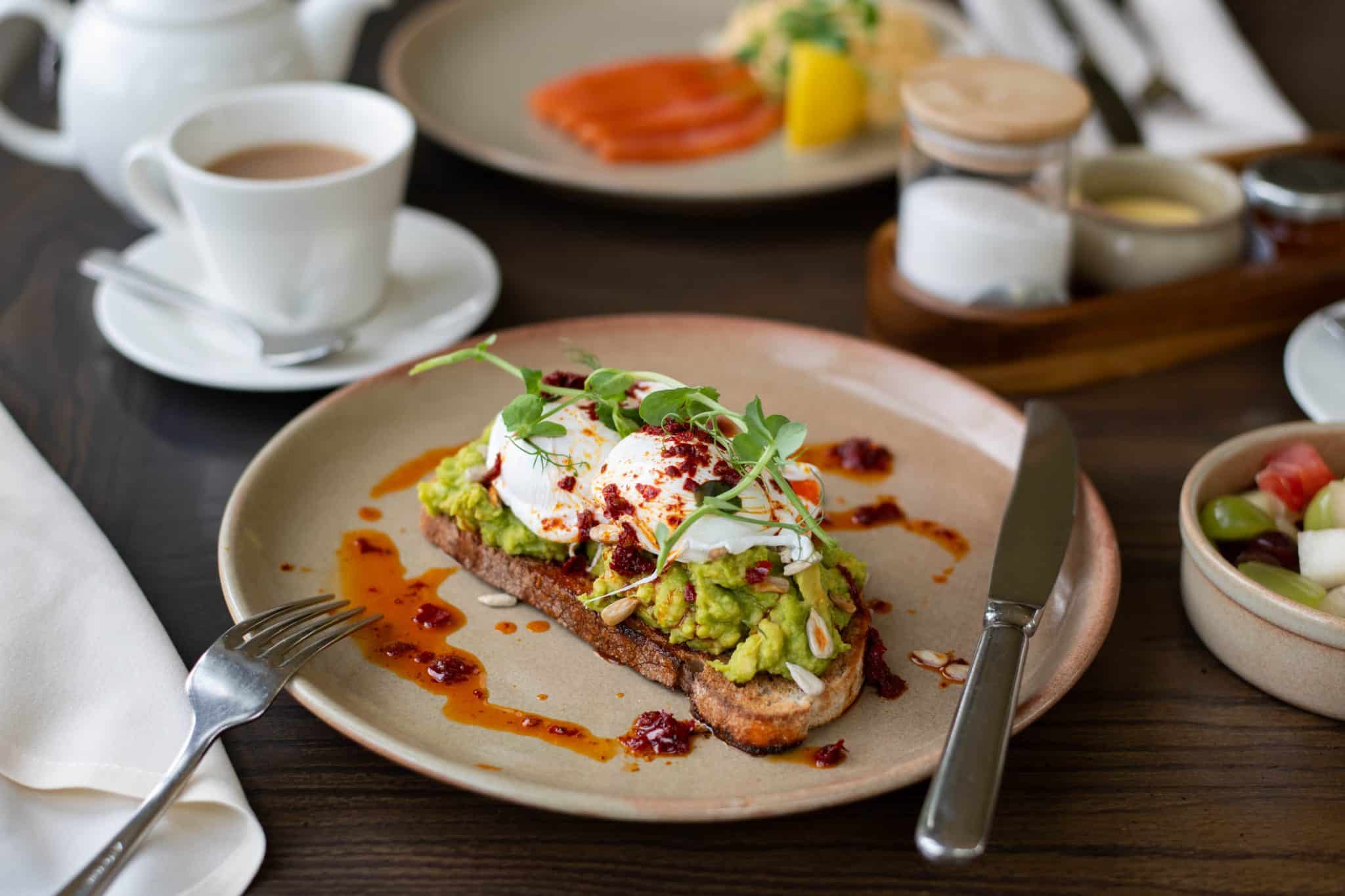 Poached eggs and avocado on toast with other breakfast items in the background