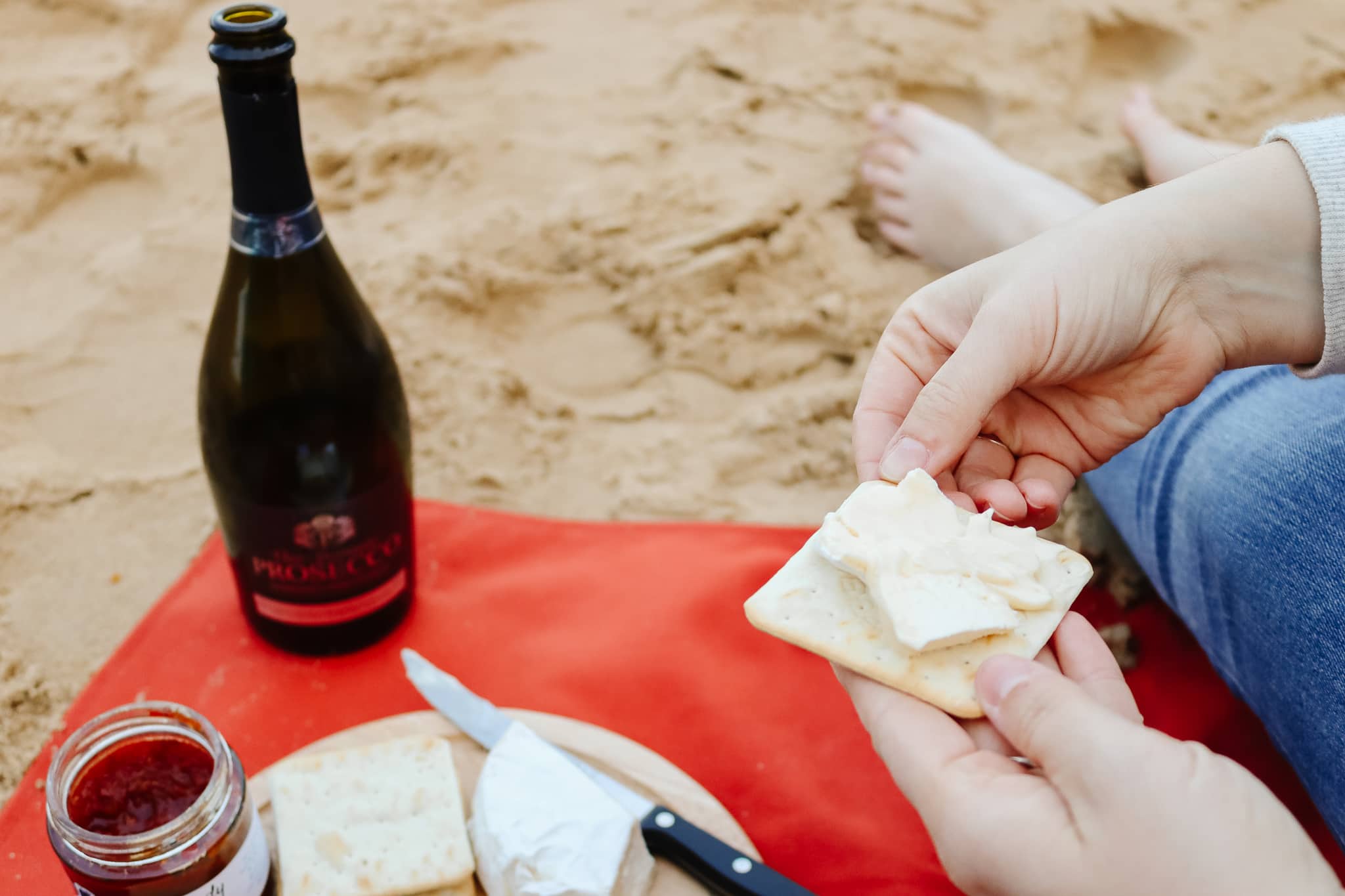 A small cheese board and an open bottle of Prosecco on a blanket on the beach