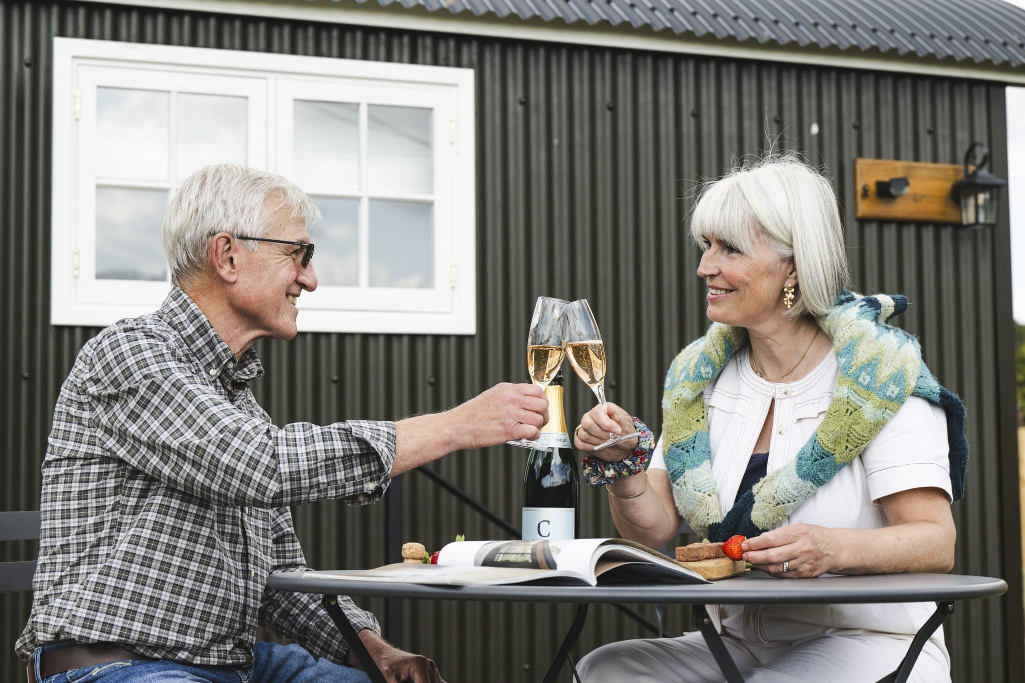 An older couple look lovingly at each other as they 'cheers' with glasses of champagne outside a shepherd hut at The Victoria