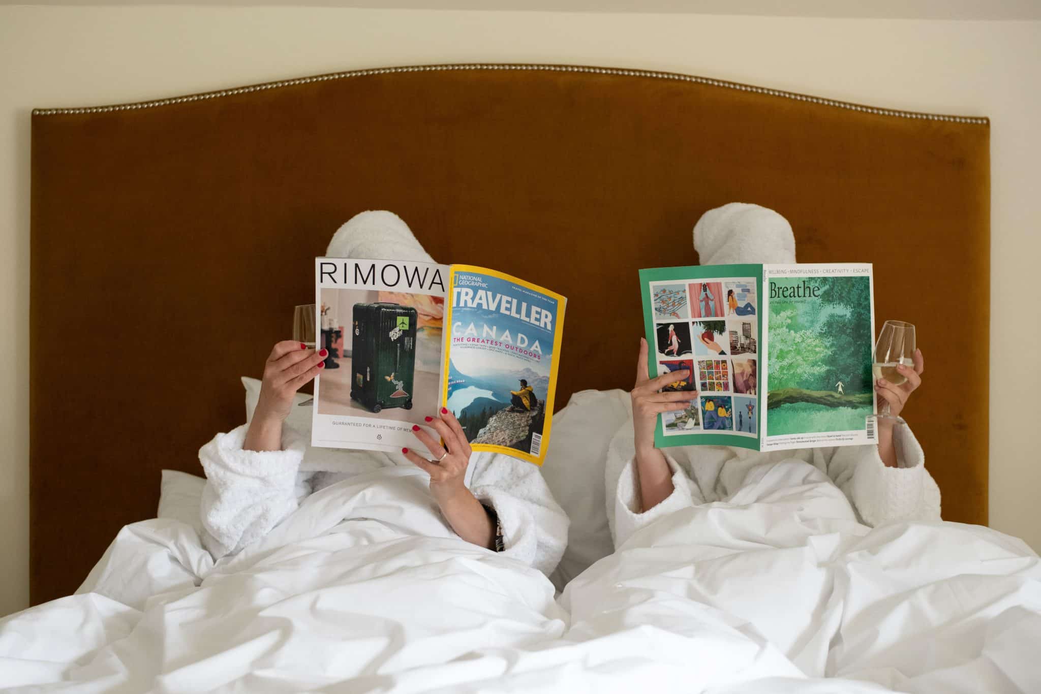 Two women in bath robes and hair towels read magazines in bed