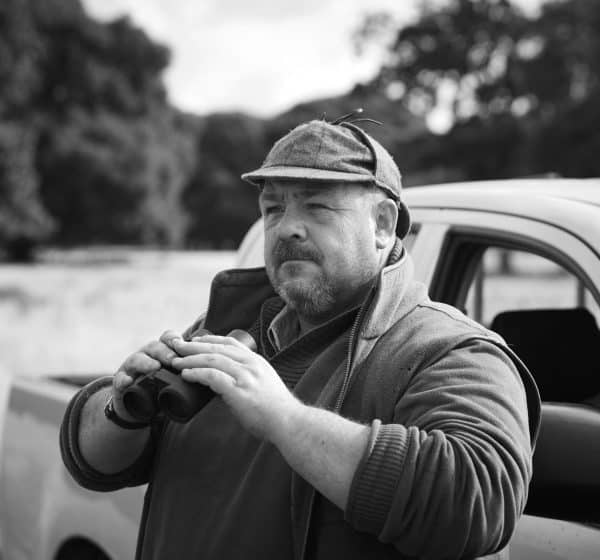A white man in country sports clothing holding a pair of binoculars.